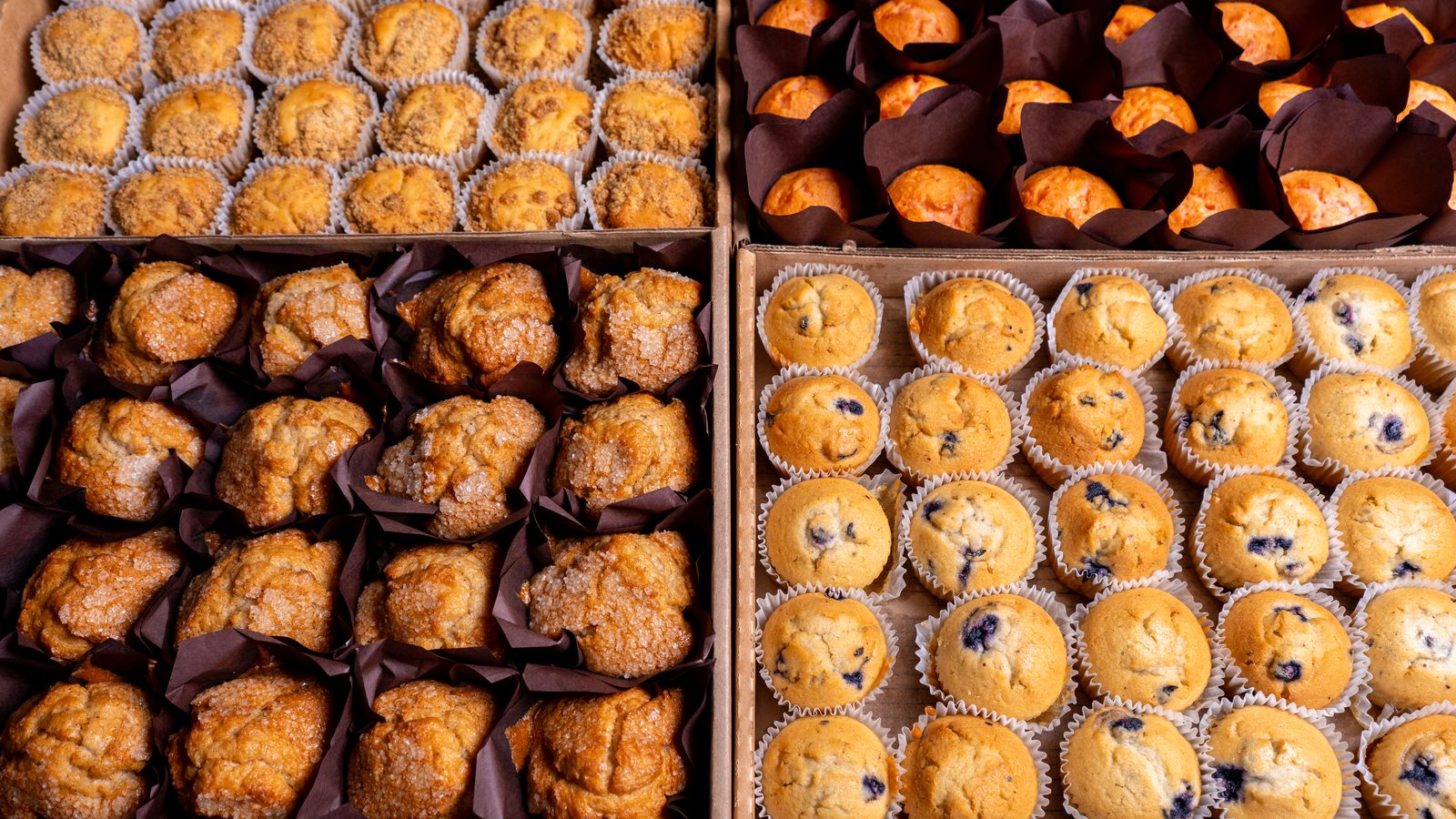 Bulk trays of coffee cake, sugar, pumpkin, and blueberry muffins for food service.